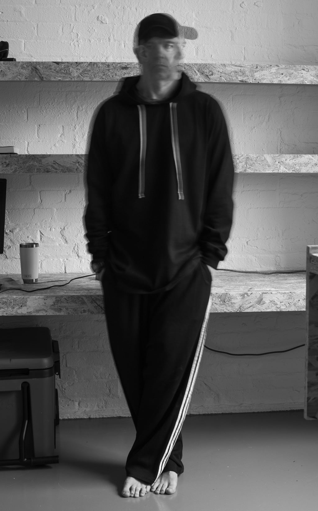 Black and white photo of person wearing a black hoodie and trousers with white stripes in workspace.